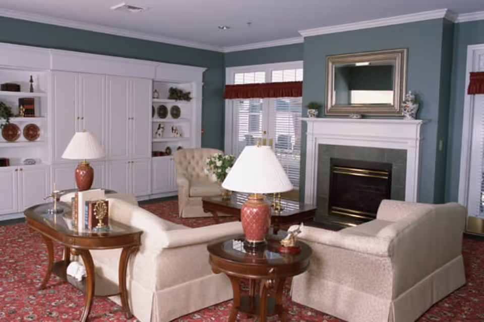 A cozy living room with beige upholstered sofas and armchairs arranged around a wooden coffee table and side tables with red lamps. The room features a fireplace with a large mirror above it, built-in white shelves with decorative items, and large windows with red valances. The walls are painted green and the floor is covered with a red patterned carpet.