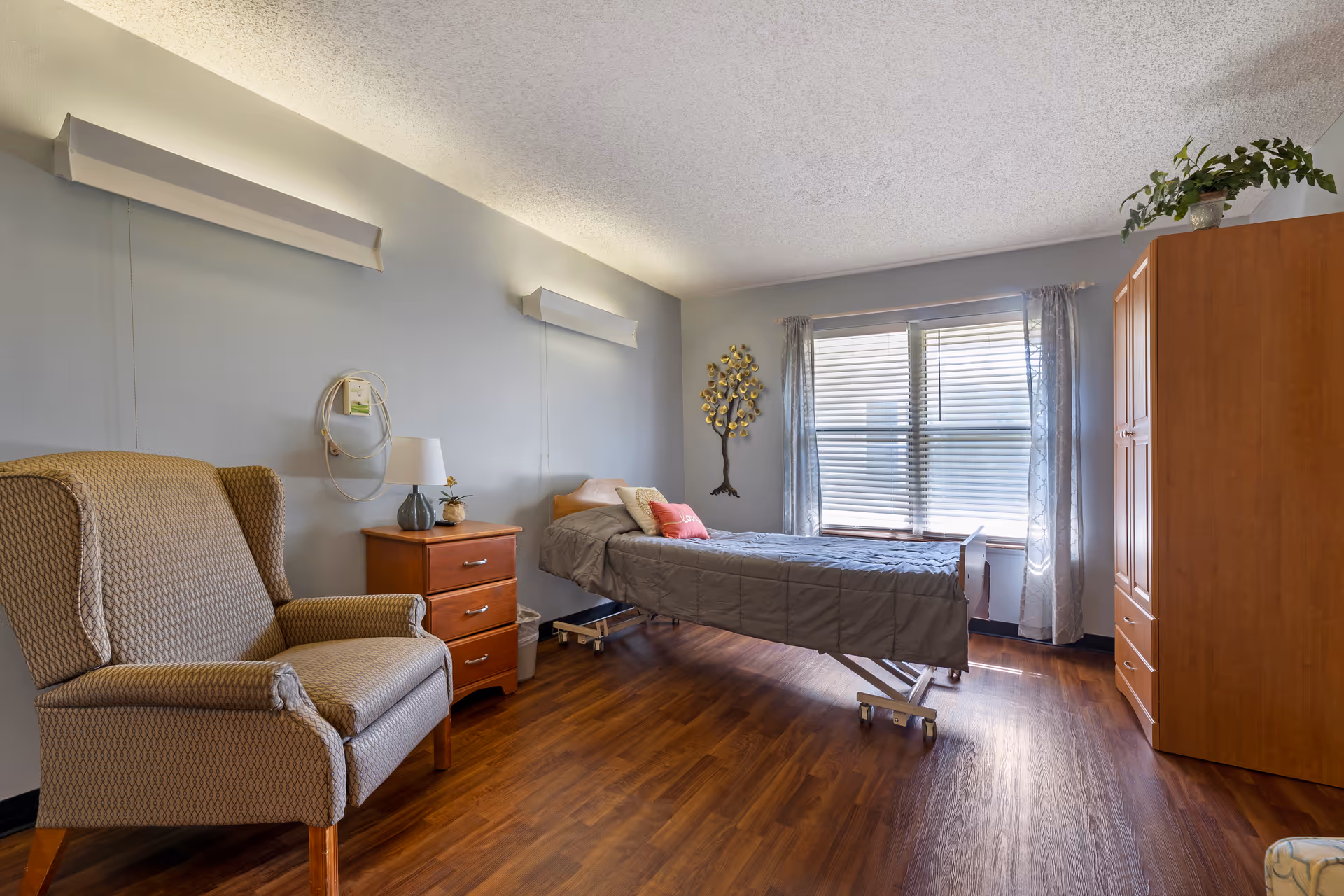 A well-lit bedroom in a senior living facility featuring a single adjustable bed with gray bedding, a wooden nightstand with a lamp and small plant, a comfortable upholstered armchair, a wooden wardrobe, and a window with blinds and sheer curtains. The room has wood flooring and light gray walls with decorative wall art.