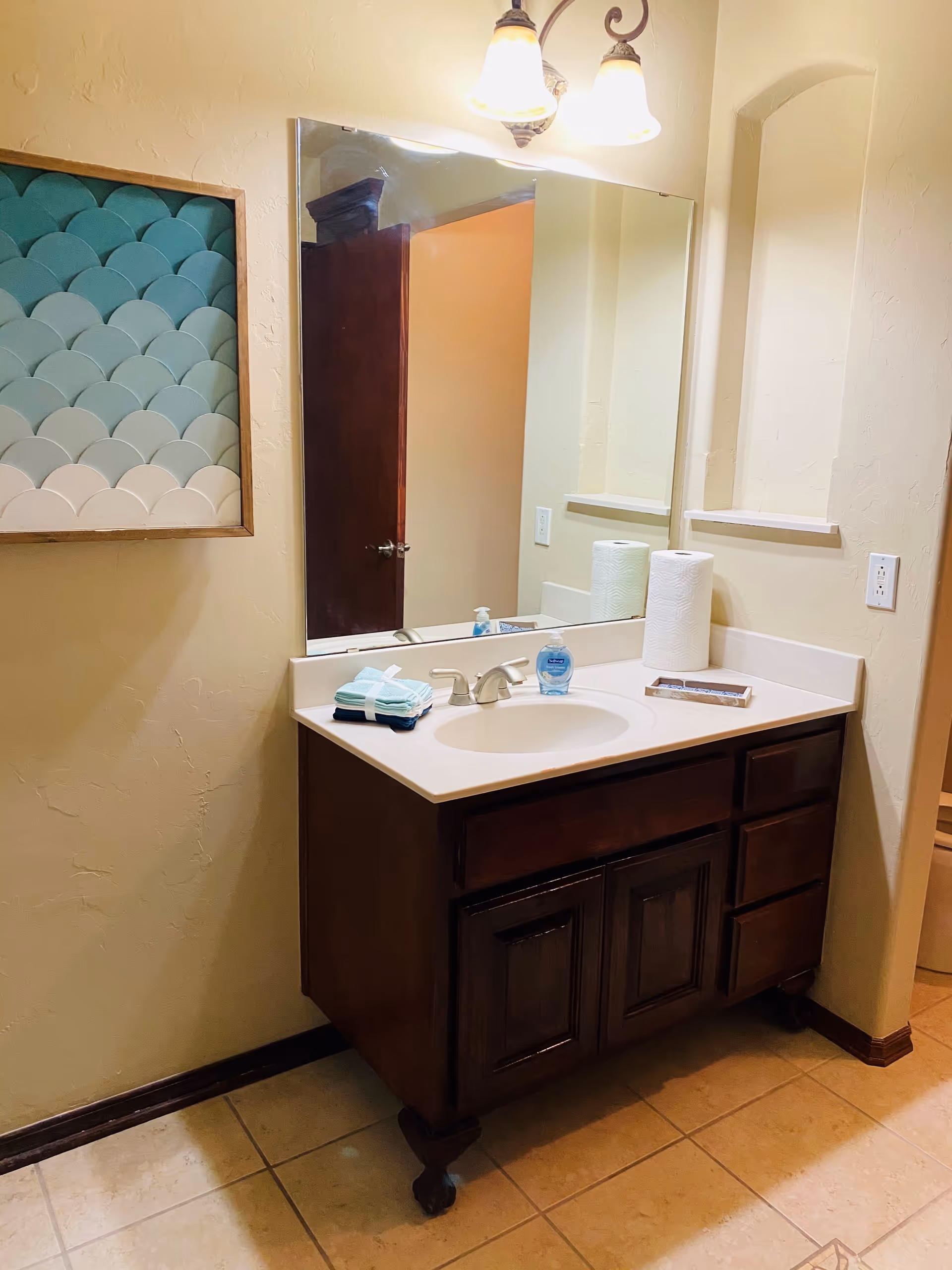 Bathroom vanity with a sink, mirror, overhead lights, soap and paper towels, and decorative wall art.