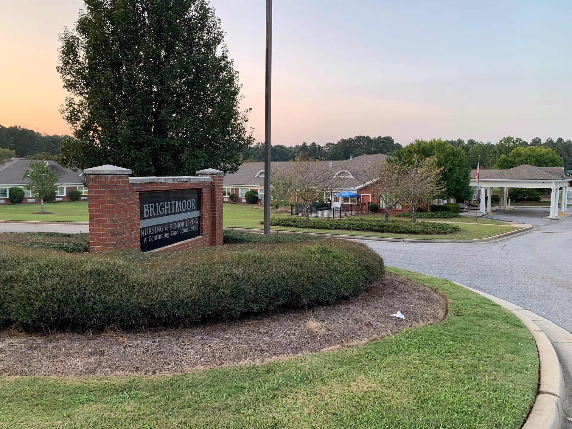 Entrance to Brightmoor Nursing & Senior Living facility with a brick sign surrounded by bushes and a large tree behind it. The building and driveway are visible in the background under a clear sky at sunset.