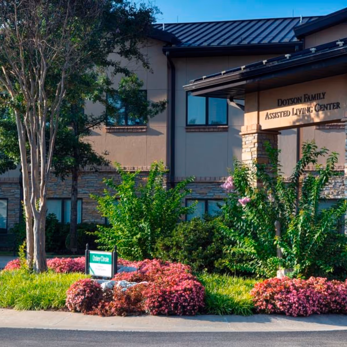 Front entrance of an assisted living building with landscaped flower beds, trees, and a sign reading "Dotson Family Assisted Living Center".