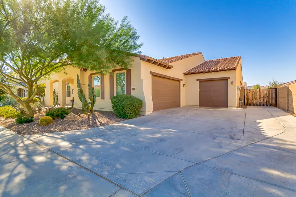 Exterior view of a single-story residential building with a beige stucco finish and brown tiled roof. The building features two brown garage doors, a driveway, and a front yard with desert landscaping including a tree, cacti, and bushes under a clear blue sky.