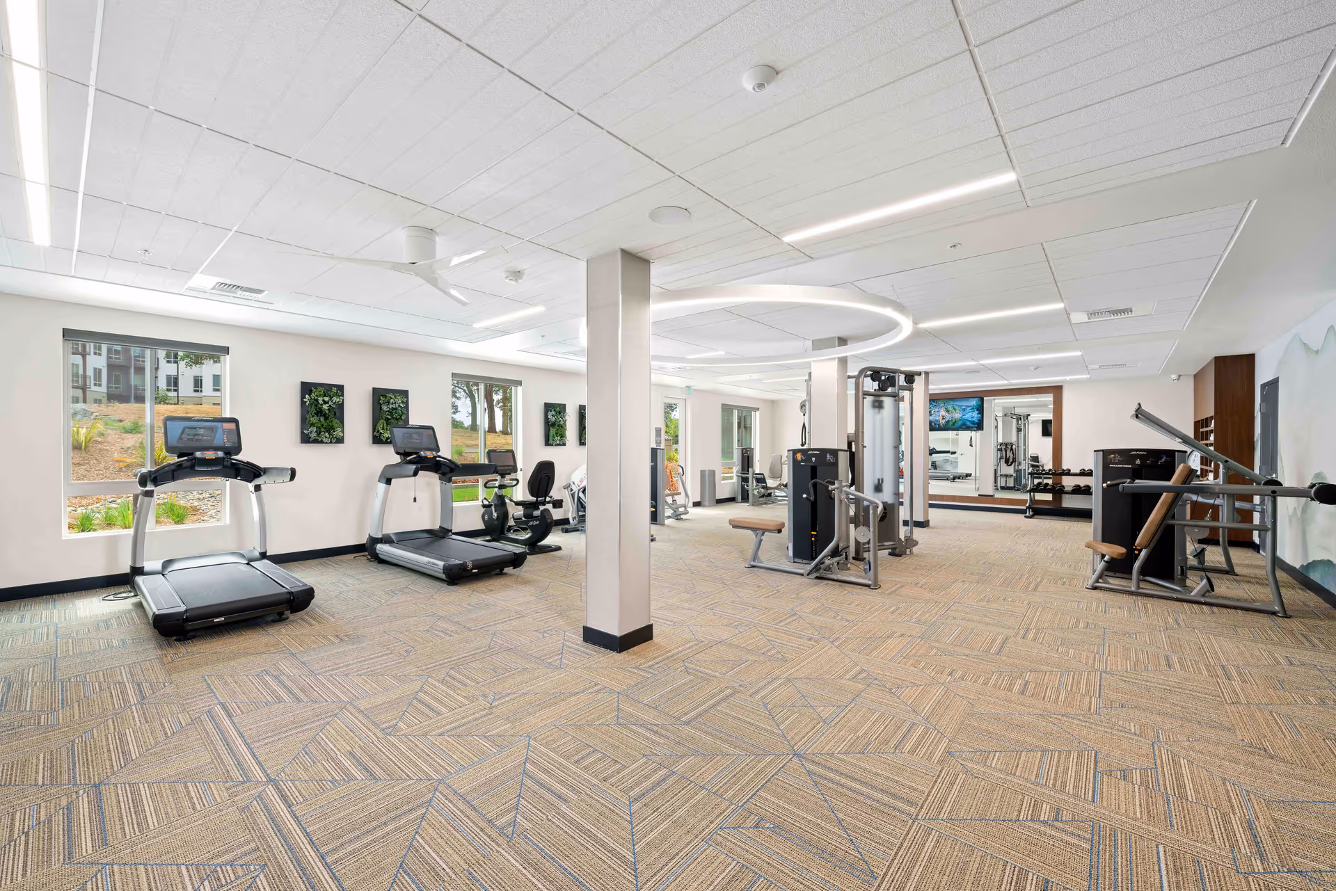 A spacious fitness room with exercise equipment including treadmills, stationary bikes, and weight machines. The room has large windows letting in natural light, modern ceiling lights, and a patterned carpet floor. There are mirrors on the far wall and some green wall art decorations.