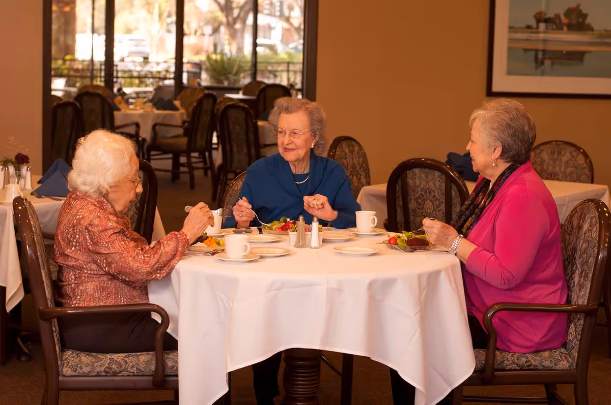 Three elderly women sitting around a round table with a white tablecloth in a dining room, eating and conversing. The room has several other tables and chairs, with large windows in the background letting in natural light.