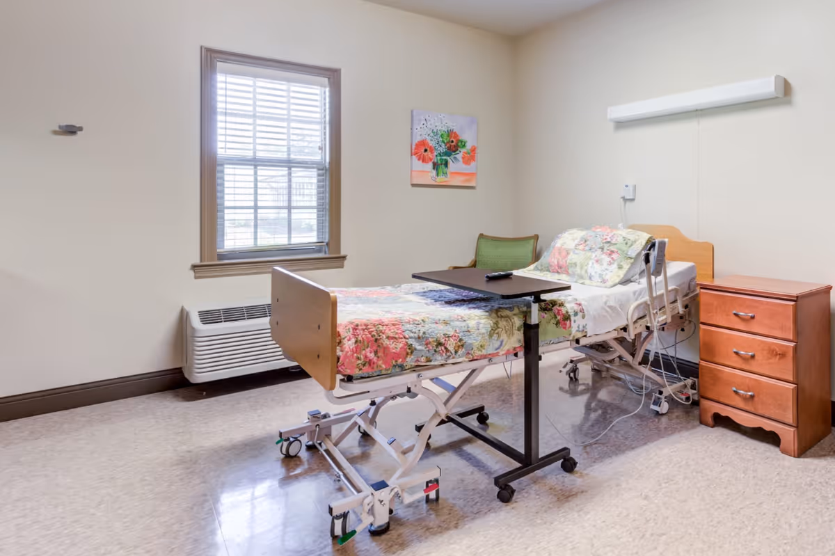 A simple, clean patient room with a hospital bed covered in floral bedding, a small wooden nightstand with three drawers, a green chair, a window with blinds, and a colorful floral painting on the wall.