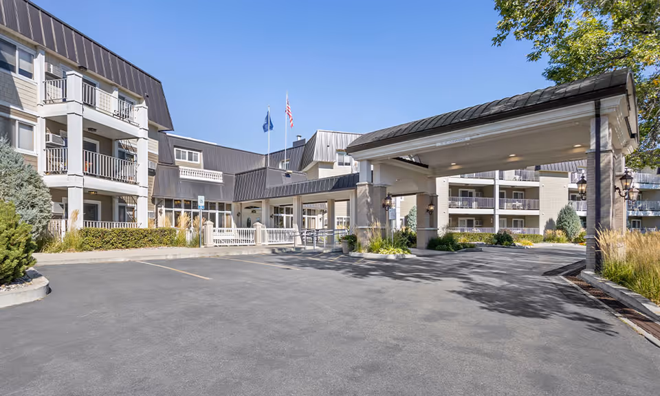 Exterior view of Westpark Village senior living facility showing a large covered entrance with columns, multiple balconies, and flags flying on flagpoles under a clear blue sky.