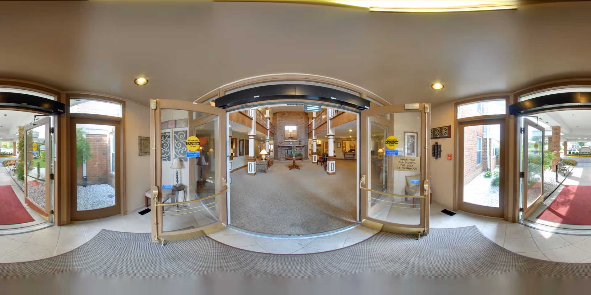 Wide-angle view of the entrance to a senior living facility lobby. Double glass doors open inward from a tiled entryway with a gray mat. Inside, a spacious carpeted lobby features a large stone fireplace at the far end, flanked by seating areas and warm lighting. Large windows and glass doors allow natural light to brighten the space. The exterior visible through the side doors shows landscaped areas with white rocks and greenery.