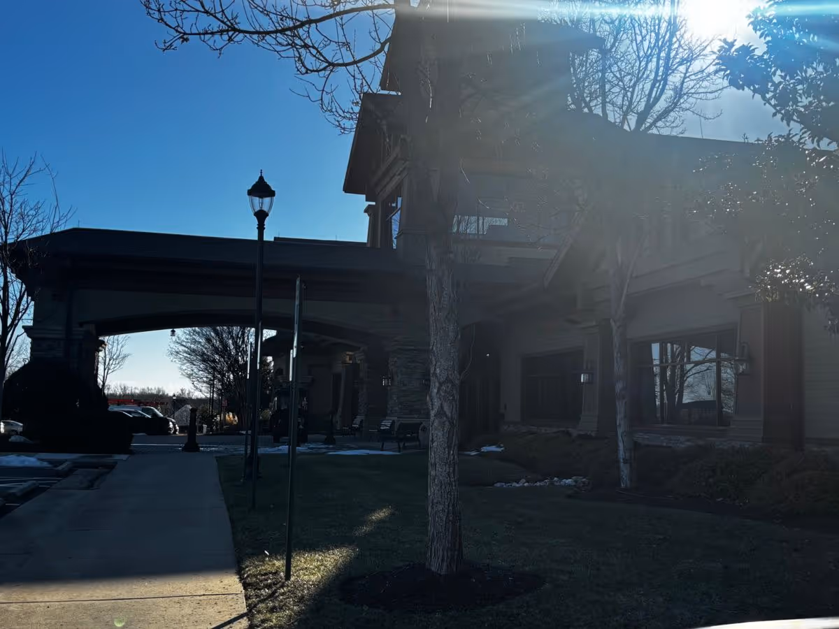 Front entrance of a senior living building with a covered porte-cochere, lamp post, sidewalk, and trees in bright sunlight.