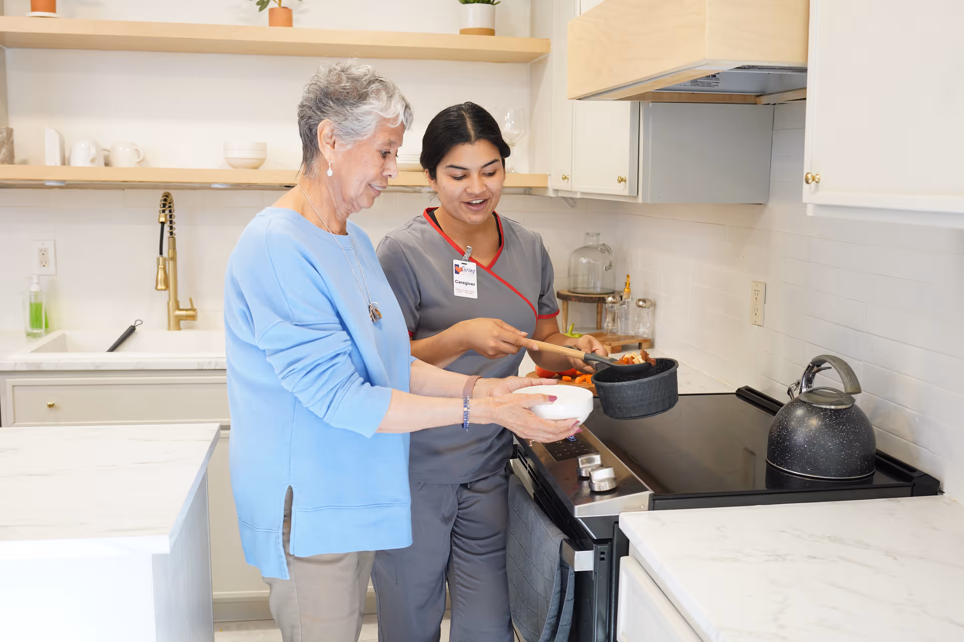 An elderly woman and a caregiver in gray scrubs are cooking together in a modern kitchen. The caregiver is holding a pot with food, while the elderly woman holds a white bowl. The kitchen has white cabinets, a black stove, a kettle, and a marble countertop.