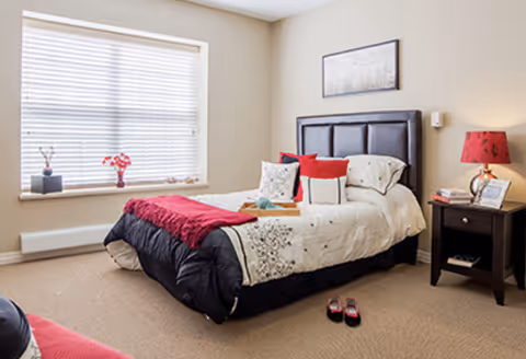 Bright bedroom with a single bed dressed in white, black, and red linens, a nightstand with a lamp, and a large window with blinds.
