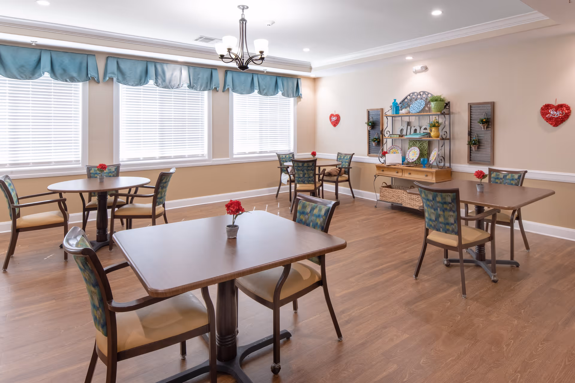 A bright and clean dining room in a senior living facility with several wooden tables and chairs arranged neatly. Each table has a small red flower centerpiece. Large windows with blue valances allow natural light to fill the room. A decorative shelving unit with plates, plants, and the word 'Love' is mounted on the far wall, which is also adorned with heart-shaped wall hangings.