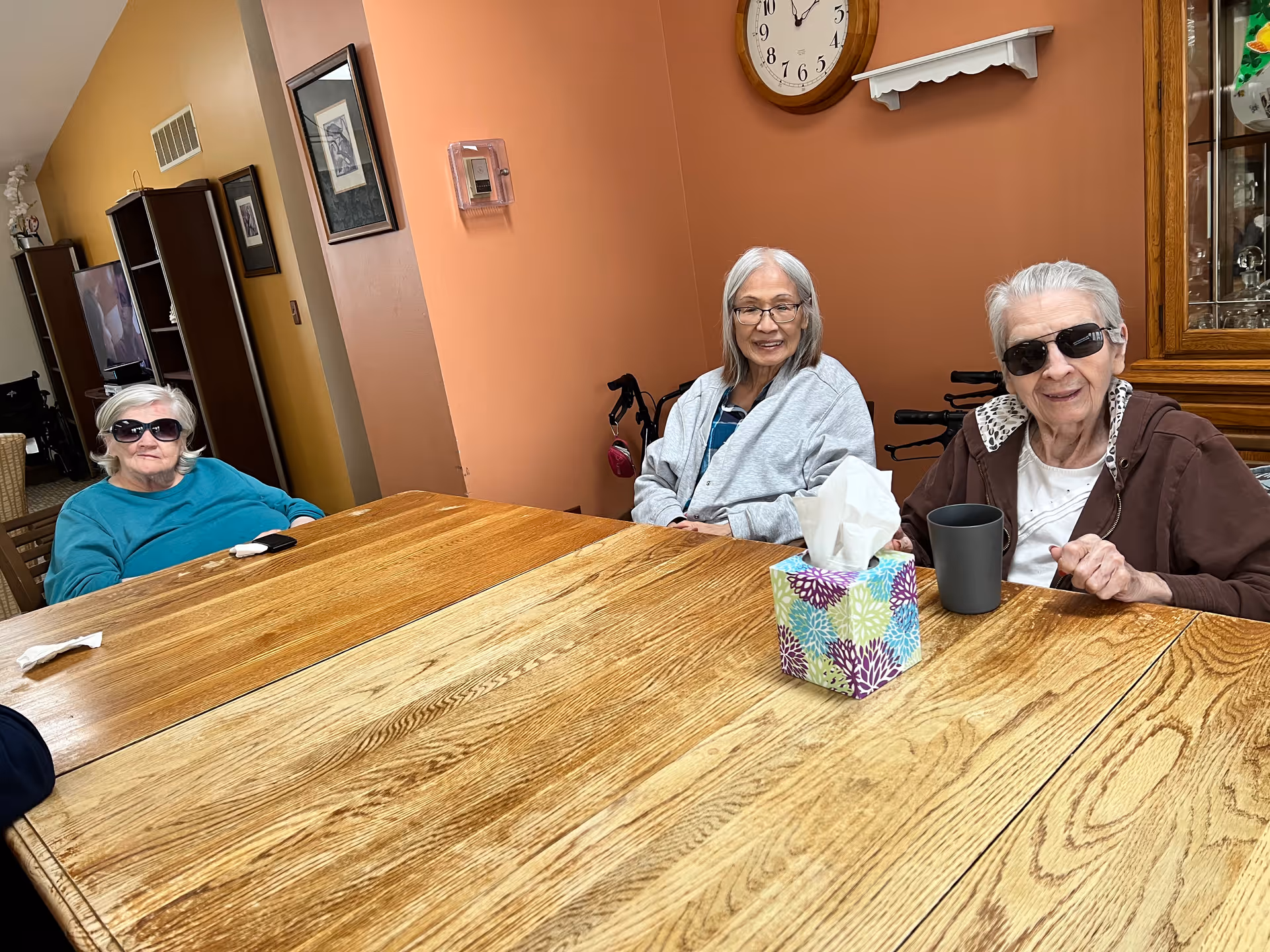 Three elderly women sitting around a wooden table in a room with orange walls. Two of the women are wearing sunglasses, and there is a tissue box and a black cup on the table. A clock is mounted on the wall behind them.
