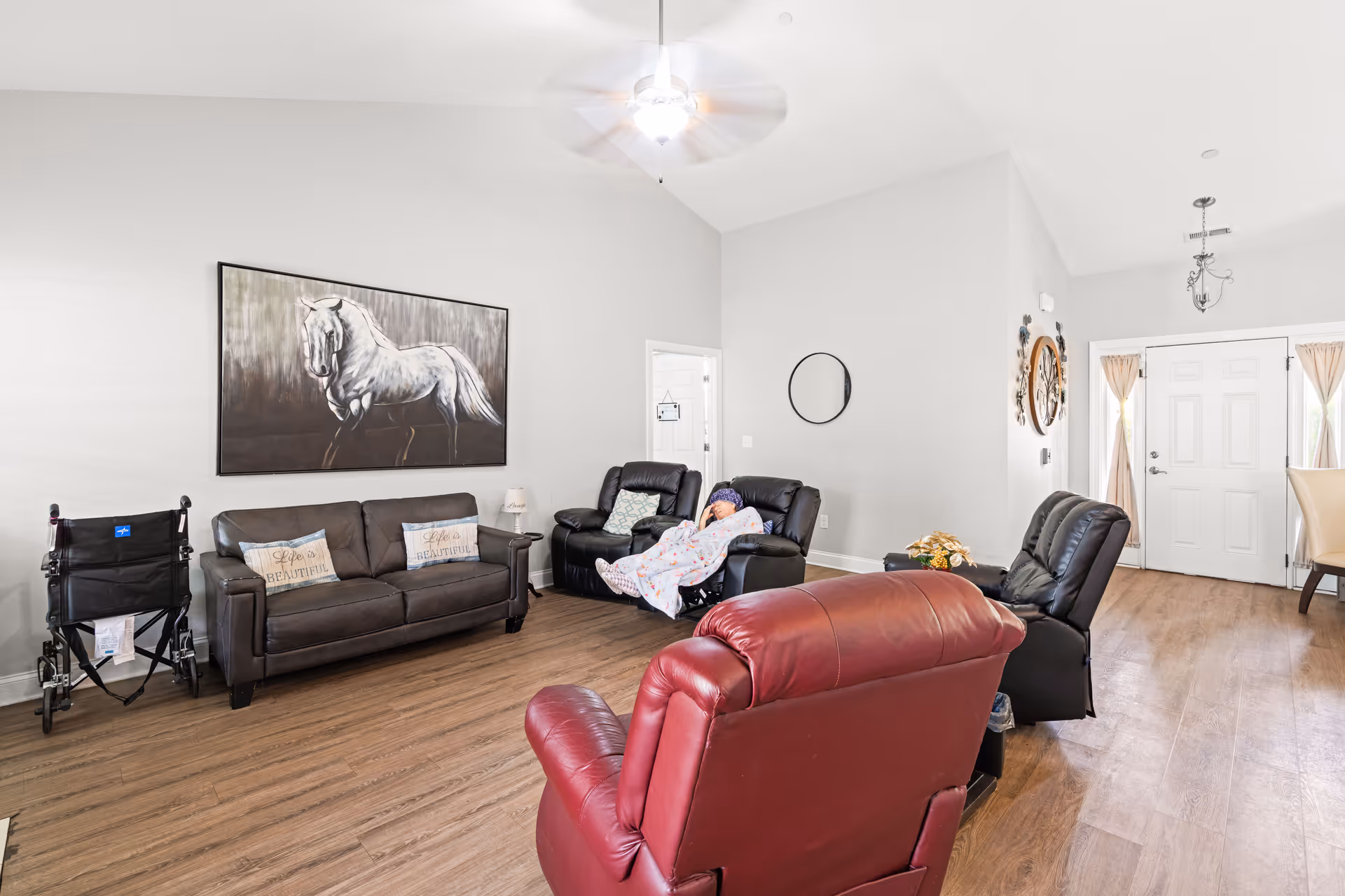 Bright assisted-living common room with sofas and recliners (one occupied by a person under a blanket), a wheelchair, a large horse painting, and the front door.