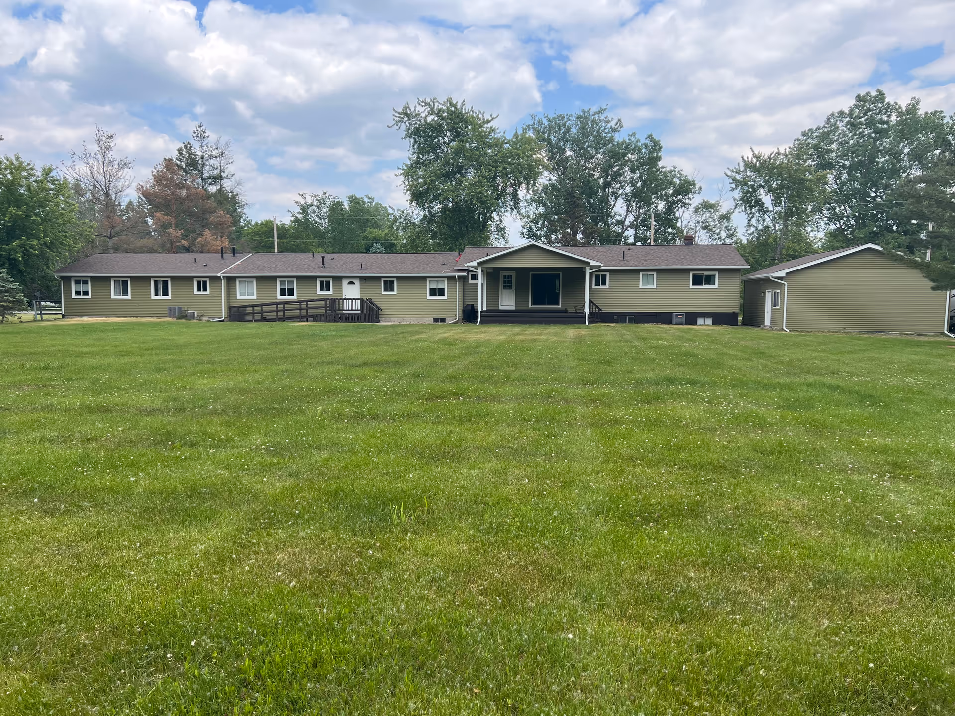 A single-story building with a covered entrance and a ramp, surrounded by a large green lawn and trees in the background under a partly cloudy sky.