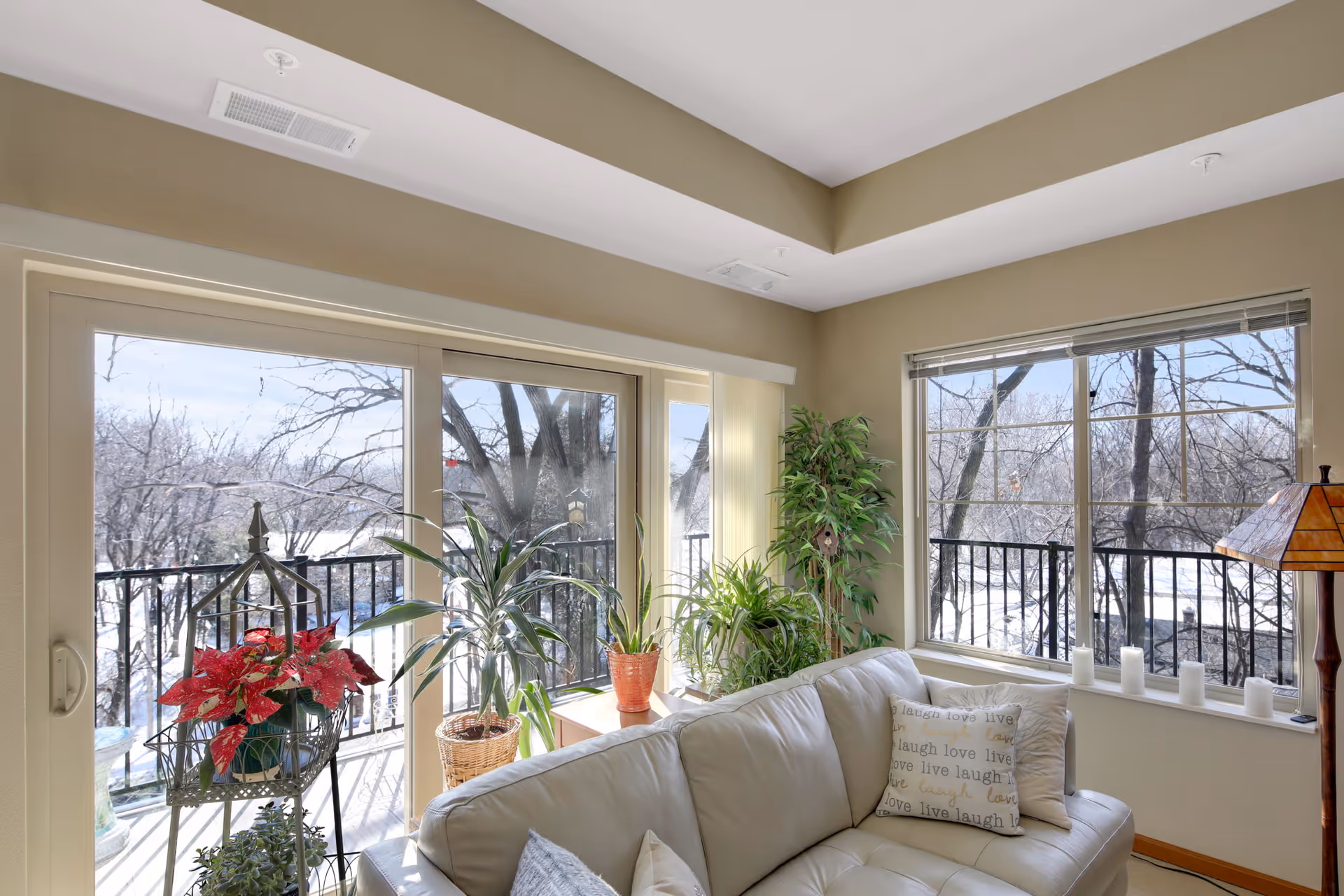 A bright living room with large windows and sliding glass doors that open to a balcony. The room features a white leather sofa with decorative pillows, several potted plants, and a floor lamp with a stained glass shade. Outside, leafless trees are visible, indicating a winter season.