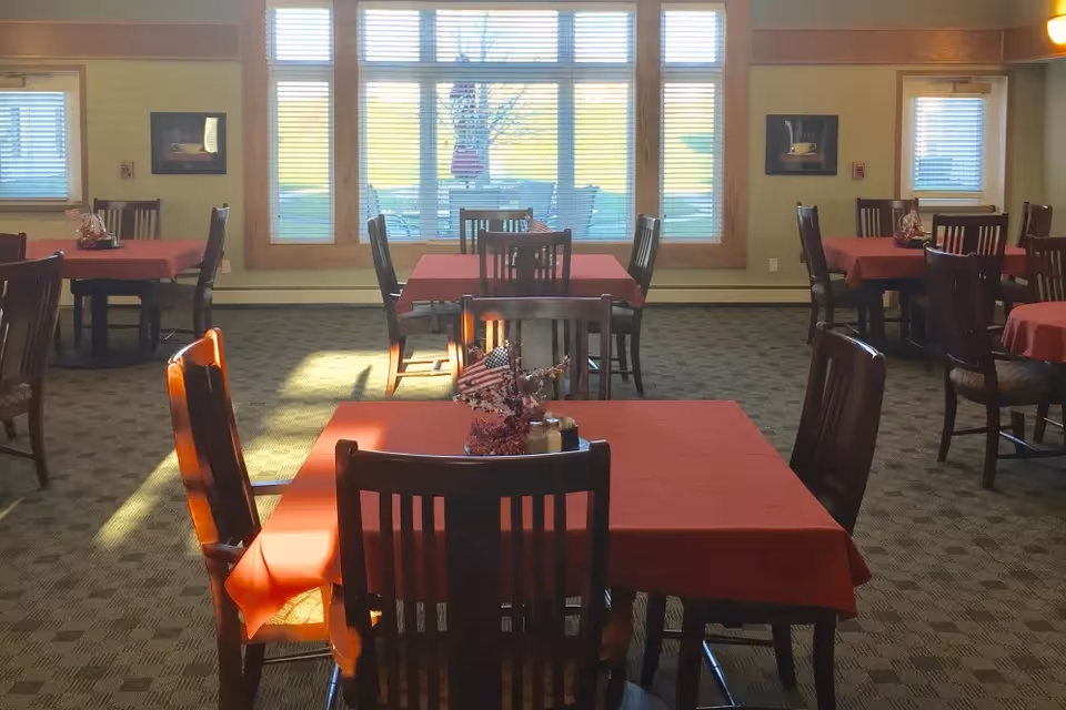 A dining room with several tables covered in red tablecloths and wooden chairs. The room has large windows letting in natural light, and there are small decorative centerpieces on the tables. The carpet has a patterned design, and framed pictures hang on the walls.