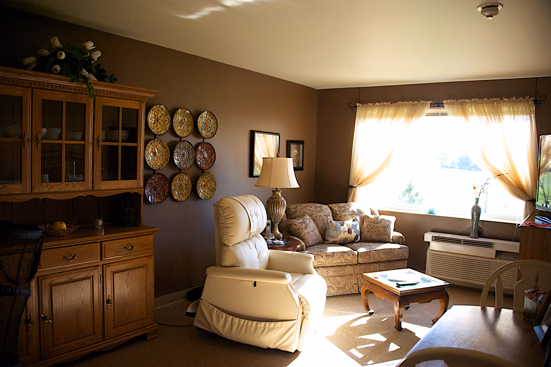 A cozy living room with a beige recliner chair, a patterned sofa with decorative pillows, a wooden coffee table, and a wooden cabinet with glass doors. The room has brown walls adorned with decorative plates and framed pictures. Light filters through sheer curtains on a large window, illuminating the space.