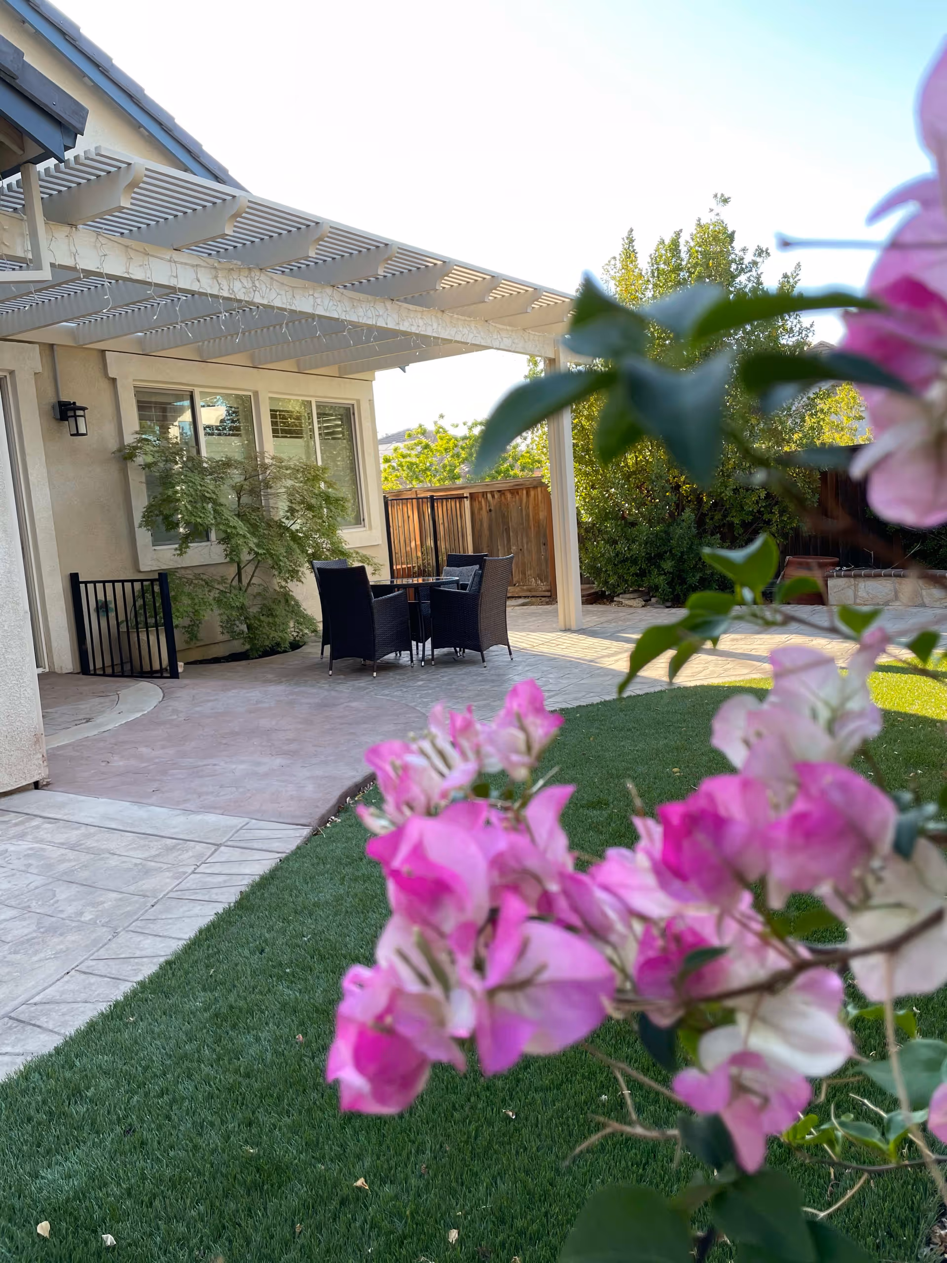 Outdoor patio area with a pergola attached to a beige building. There is a small table with four black wicker chairs on a concrete patio. In the foreground, there are pink and white flowers with green leaves. The area is surrounded by a wooden fence and greenery.