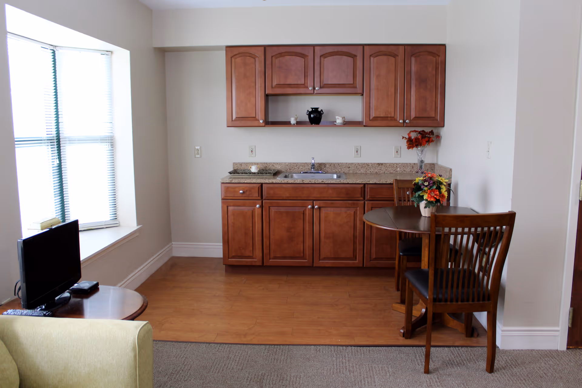 A small kitchenette area with wooden cabinets, a sink, and a granite countertop. To the right, there is a round wooden table with two chairs and a vase of flowers on top. To the left, a window with blinds lets in natural light, and a small TV sits on a round wooden side table next to a green armchair.