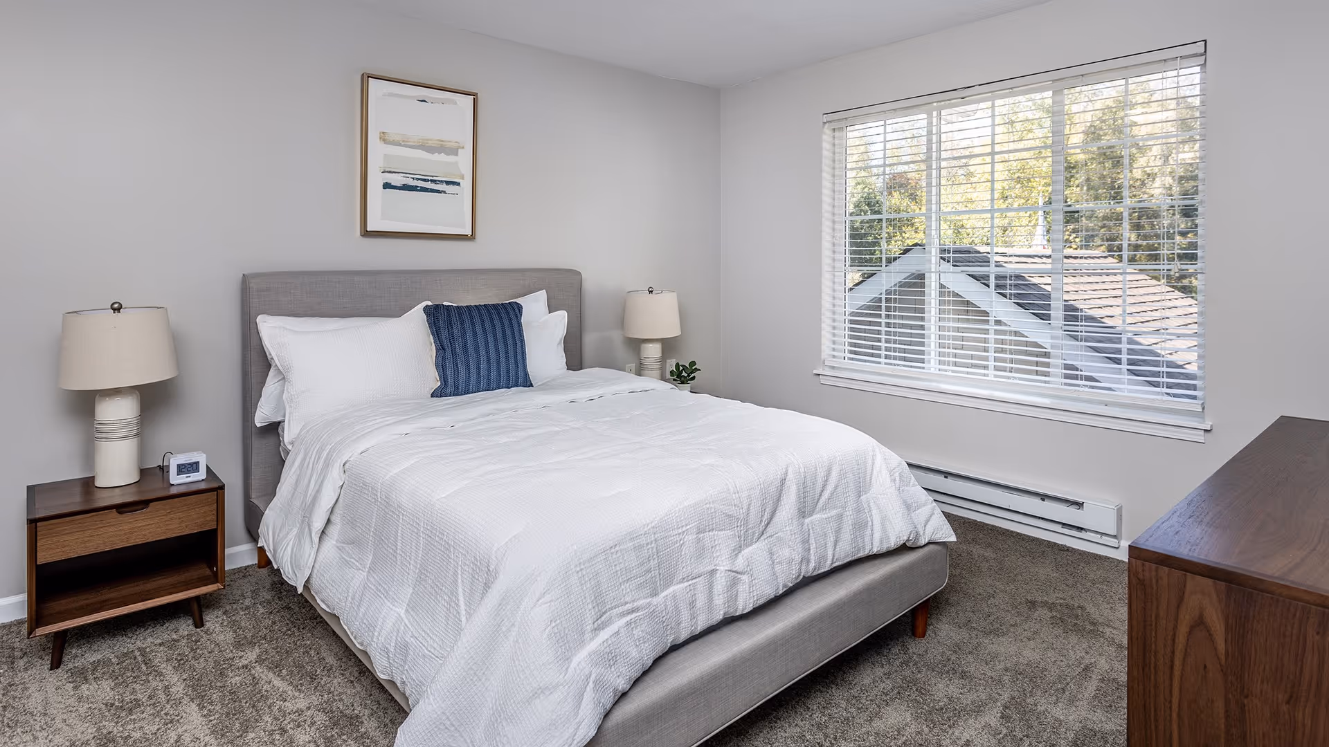 A neatly made bed with white bedding and a blue accent pillow in a bedroom. The room has a gray upholstered headboard, two bedside tables each with a lamp, a framed abstract artwork on the wall, a large window with white blinds showing a view of rooftops and trees outside, and a wooden dresser on the right side.