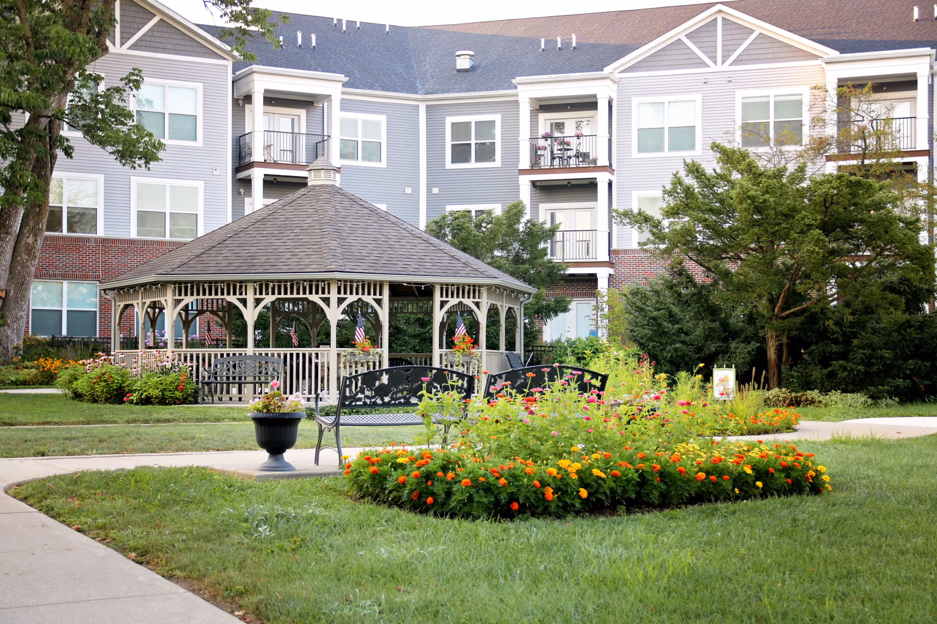 Outdoor garden area at Legacy Village featuring a large gazebo with a shingled roof, surrounded by benches and colorful flower beds. The background shows a multi-story residential building with balconies and windows, and trees providing greenery.