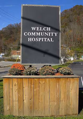 Large Welch Community Hospital sign atop a wooden planter with flowers outdoors.
