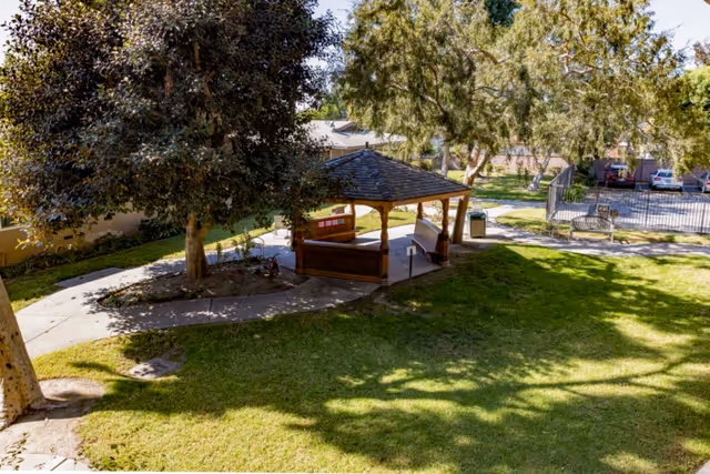 Outdoor courtyard with a wooden gazebo, trees, grassy lawn, walking paths and benches.
