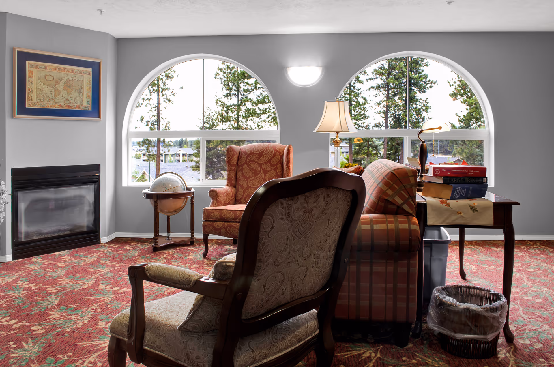A cozy living room area with patterned armchairs and a sofa arranged around a small table with books and a lamp. The room features large arched windows showing trees outside, a globe on a stand, a fireplace on the left wall, and a red floral carpet.