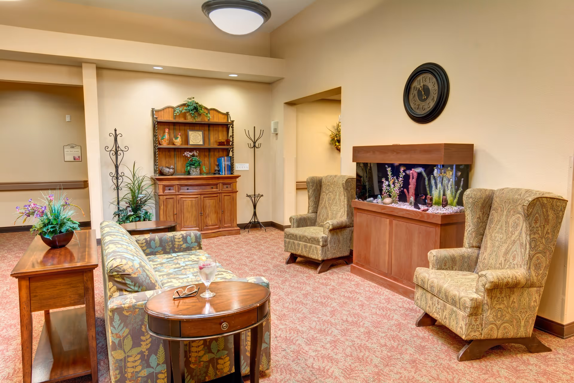 A cozy sitting area in a memory care residence featuring two patterned armchairs, a floral-patterned sofa, a wooden side table with a glass of water and eyeglasses, a wooden cabinet with decorative items, a coat rack, and a large fish tank beneath a wall clock. The room has warm beige walls and carpeted flooring with a floral pattern.