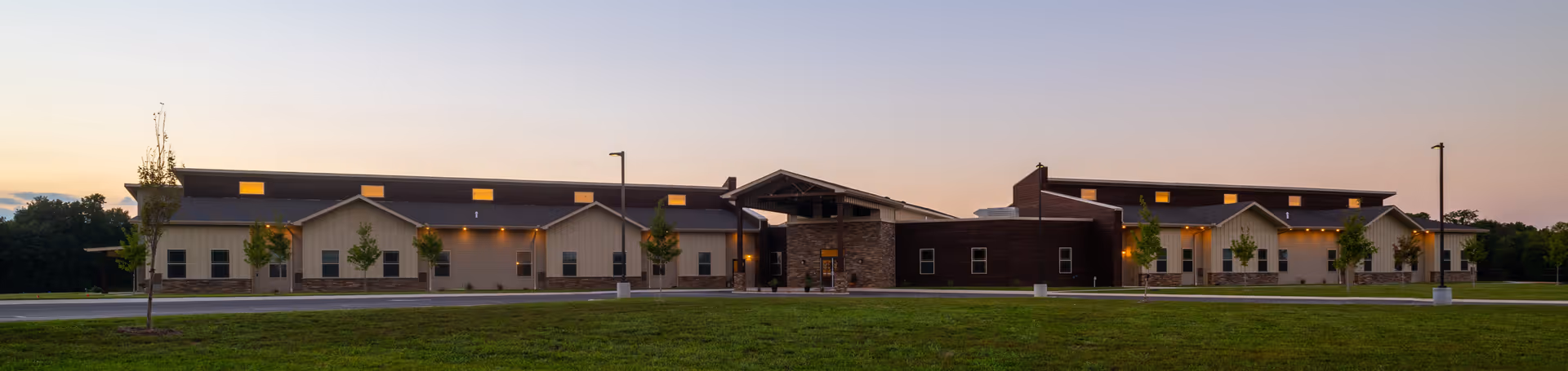 Wide exterior view of Webwood Assisted Living facility at dusk, showing a modern single-story building with multiple windows, stone and wood facade, small trees planted in front, and a well-maintained grassy area.