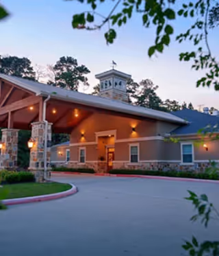 Exterior view of Village Green Memory Care Community Tomball building at dusk with a covered entrance supported by stone pillars and surrounded by trees and landscaping.