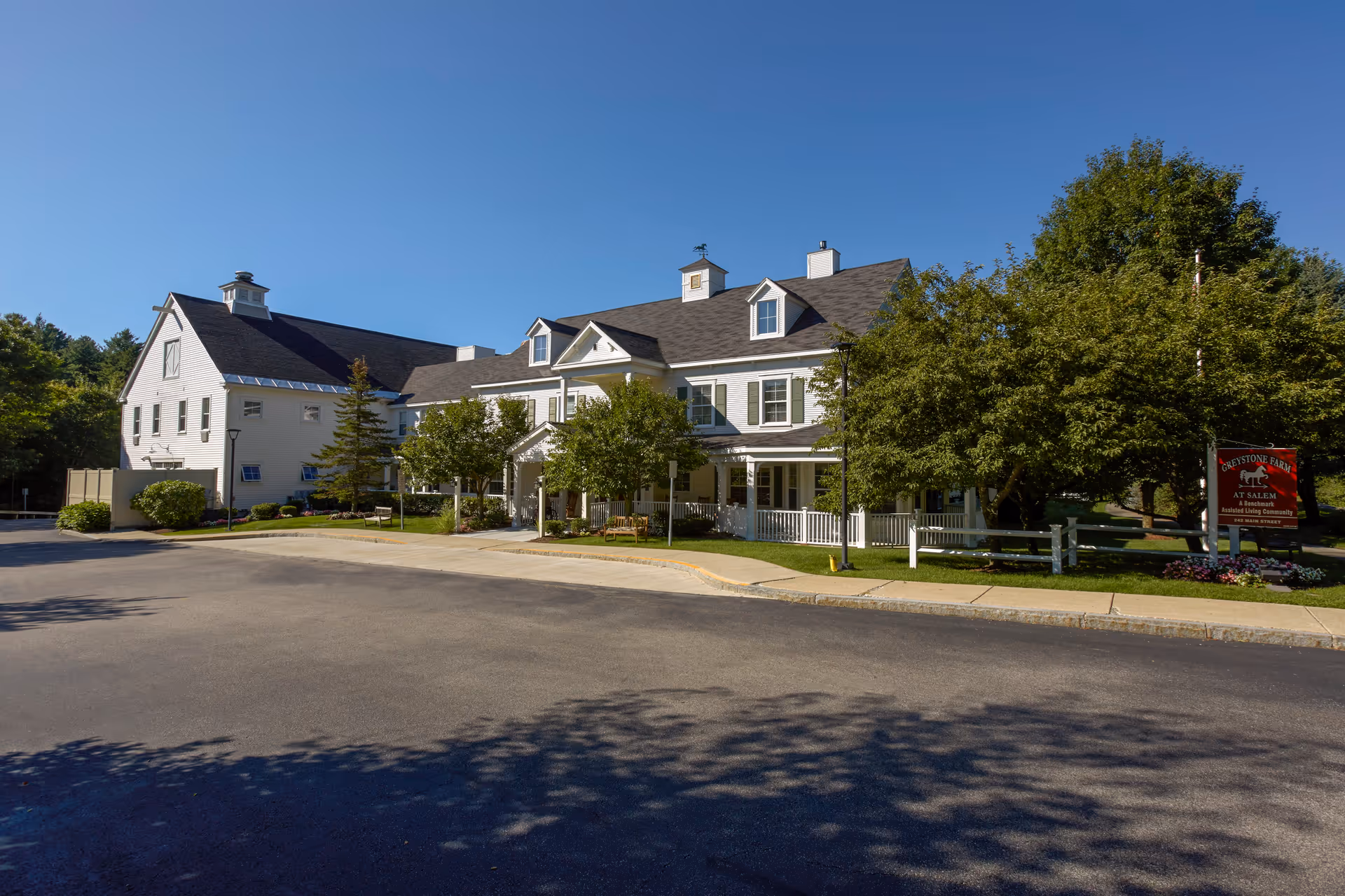 Exterior view of Greystone Farm at Salem, a large white two-story building with a porch, surrounded by trees and greenery under a clear blue sky.