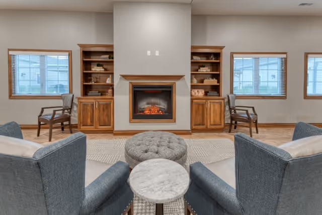A cozy senior living common area featuring a central electric fireplace flanked by wooden built-in shelves and cabinets. Two windows with wooden blinds are on either side of the fireplace. In front of the fireplace are two blue upholstered armchairs with a small round marble-top table and a tufted round ottoman between them. Two wooden chairs are positioned near the windows.
