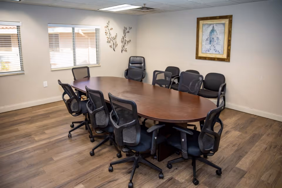 A conference room with a large oval wooden table surrounded by eight black mesh office chairs on wheels. There are three additional black chairs without wheels against the wall. The room has wooden flooring, two windows with blinds, a framed picture on the wall, and decorative wall art.