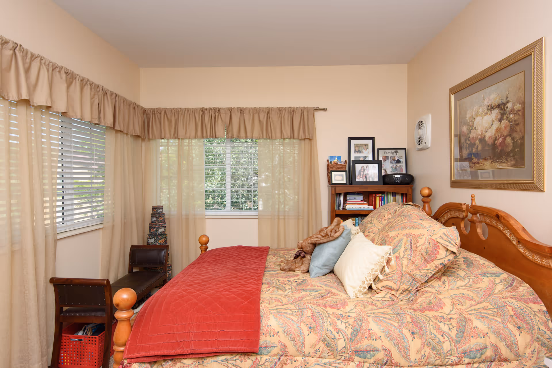 A cozy bedroom with a wooden bed frame, patterned bedspread, and multiple pillows including a red quilt. There are two windows with beige curtains allowing natural light to enter. A small wooden bench is placed near the windows, and a bookshelf with framed photos and books is against the wall. A floral painting hangs above the bed.