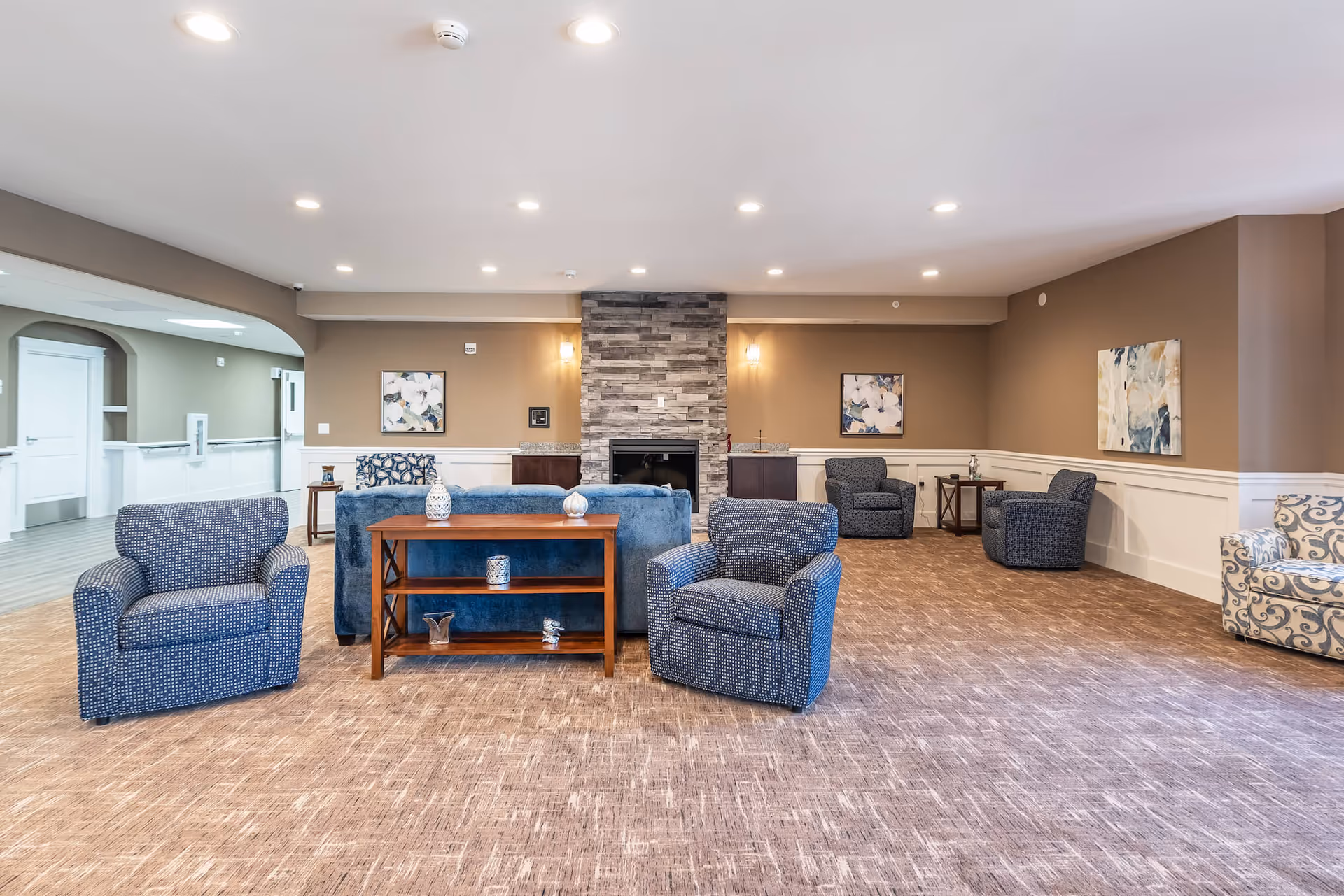 Spacious senior living common room with multiple upholstered chairs and sofas arranged around a central console table and a stone fireplace.