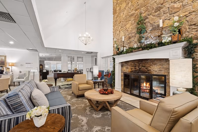A cozy living room area with a stone fireplace featuring a lit fire, decorated with candles and greenery on the mantel. The room has a high ceiling with a chandelier, a striped sofa with pillows, two beige armchairs, a round wooden coffee table with a decorative centerpiece, and a side table with a white flower arrangement. In the background, there is a grand piano and additional seating areas.