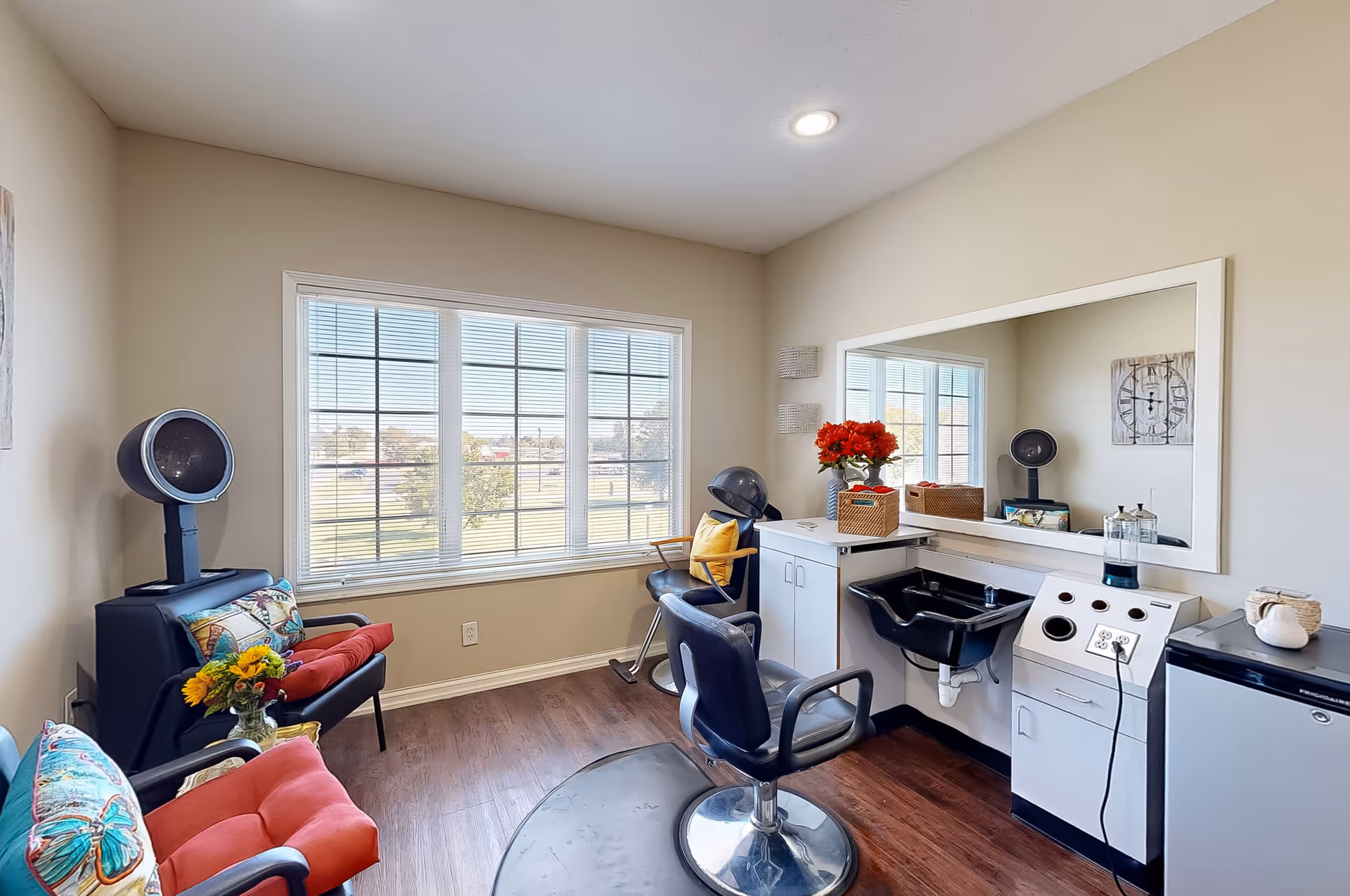 A bright and clean hair salon room with large window letting in natural light. The room features a black salon chair in front of a large mirror, a black hair washing sink, a hair dryer chair, and two red cushioned chairs with colorful pillows. There are flowers on a small table and a cabinet with hair care equipment.
