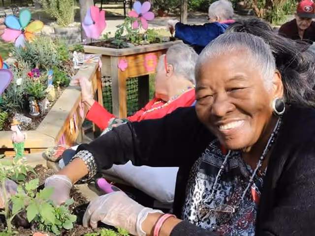 A smiling elderly woman wearing gloves is gardening in an outdoor raised garden bed decorated with colorful pinwheels. Other elderly individuals are also engaged in gardening activities in the background.