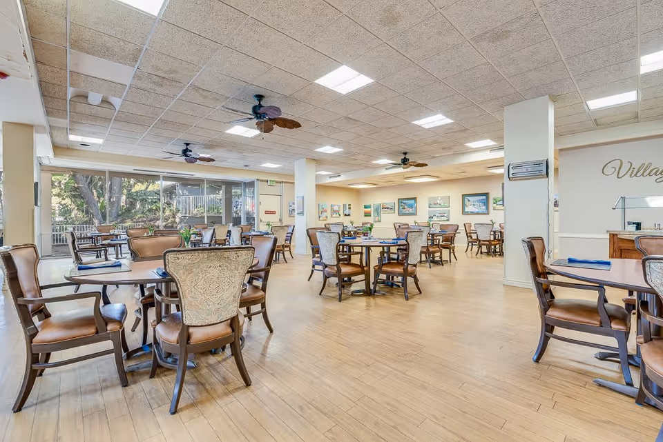 A spacious dining room with multiple round tables and cushioned chairs arranged neatly. The room has large windows letting in natural light and ceiling fans. The walls are decorated with framed pictures, and the floor is wooden. The word 'Village' is partially visible on one wall.