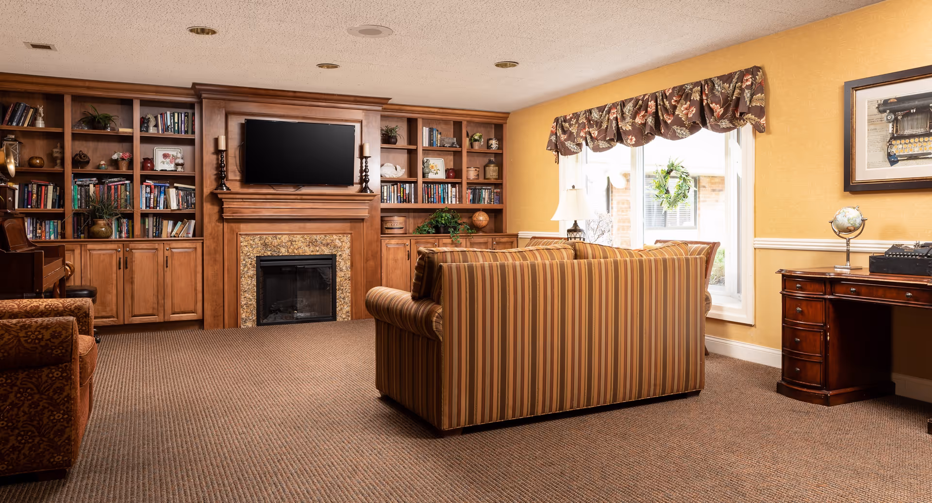 A cozy living room with a striped sofa facing a fireplace with a mounted flat-screen TV above it. The fireplace is surrounded by built-in wooden bookshelves filled with books and decorative items. To the right, there is a window with floral valance curtains, a table lamp, and a wooden desk with a globe and a vintage typewriter. The room has warm yellow walls and carpeted flooring.