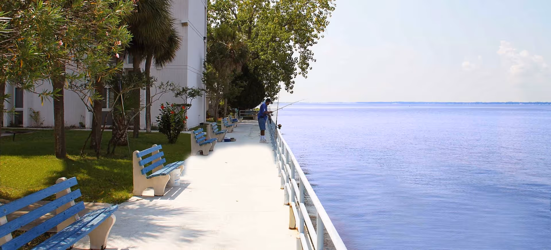 A waterfront walkway with blue benches along the left side and a white railing on the right side overlooking a large body of water. Trees and greenery are visible near the building on the left, and a person is fishing near the railing in the distance.
