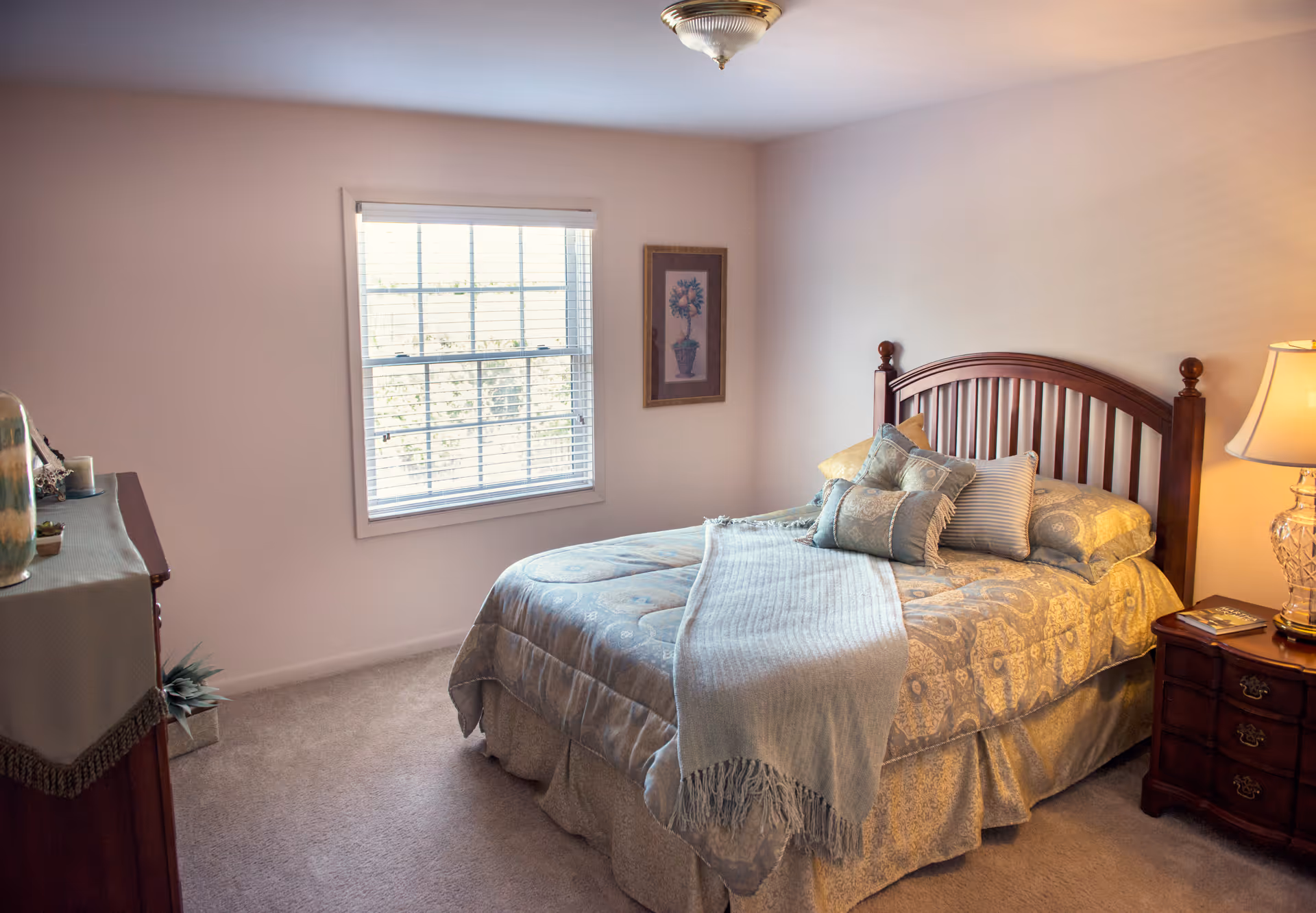 A cozy bedroom with a neatly made bed featuring patterned bedding and multiple pillows. There is a wooden headboard, a nightstand with a lamp and a book, a window with blinds letting in natural light, a framed floral picture on the wall, and a dresser with decorative items on the left side.