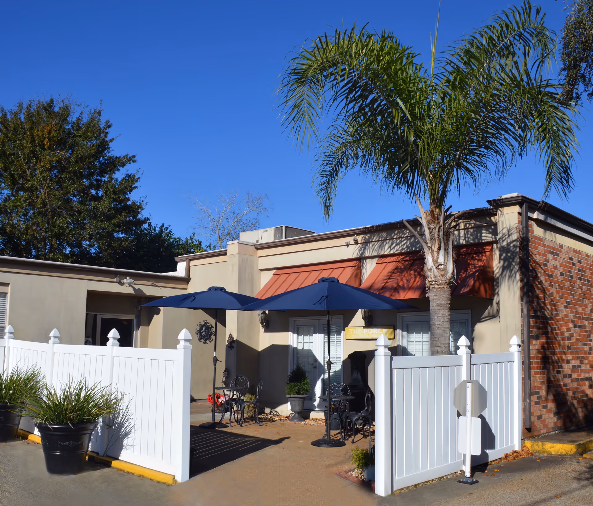 Outdoor patio area at St Anthony Community Care Center with two blue umbrellas shading black metal tables and chairs, surrounded by a white fence, a palm tree, and some potted plants, under a clear blue sky.