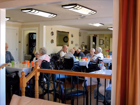 A group of elderly people, some in wheelchairs, sitting around tables in a well-lit dining area with white tablecloths and colorful cups. The room has beige walls, ceiling lights, and framed decorations on the walls.