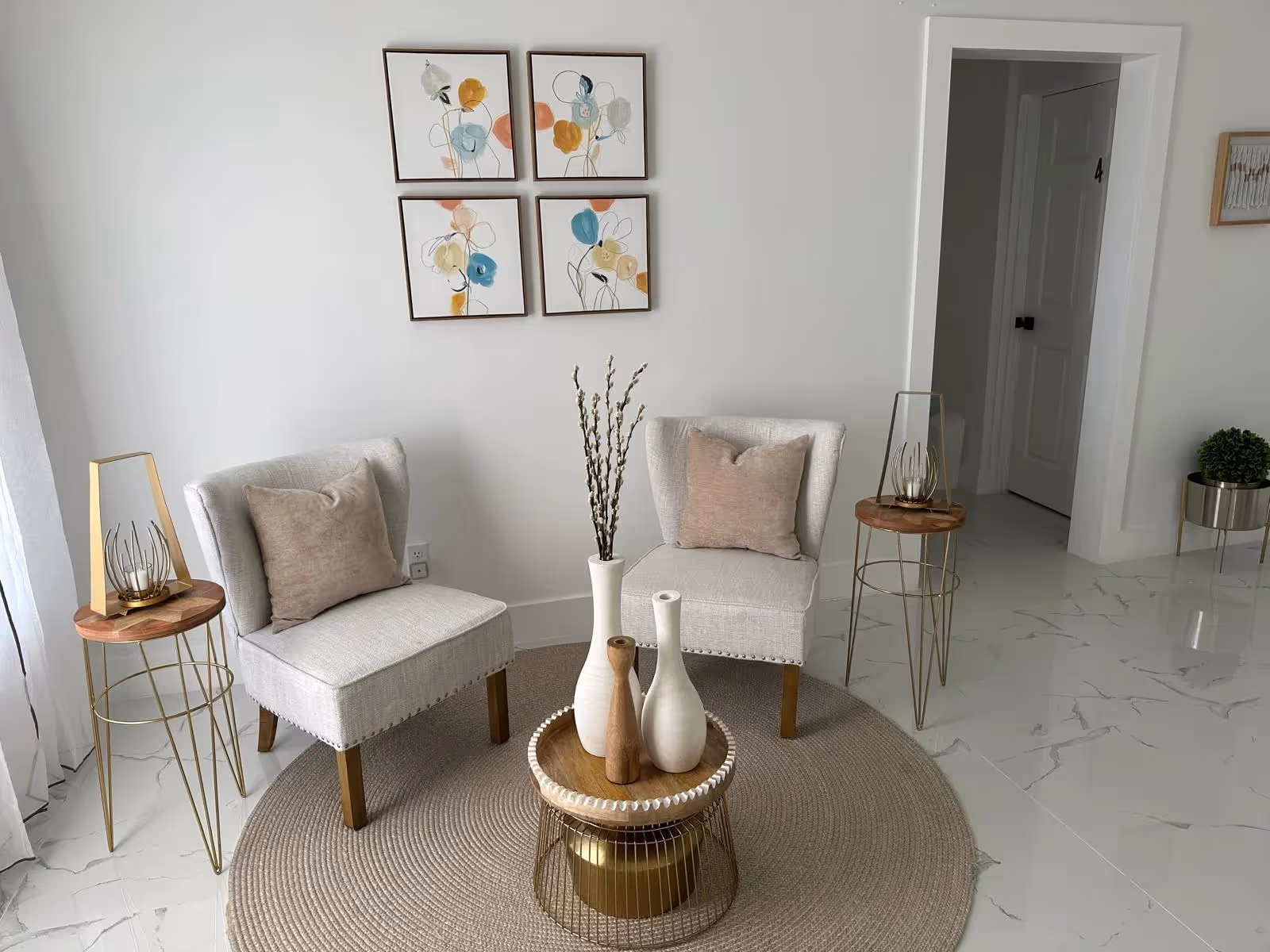 A bright, modern living room seating area with two beige chairs, a round woven rug, and abstract wall art.