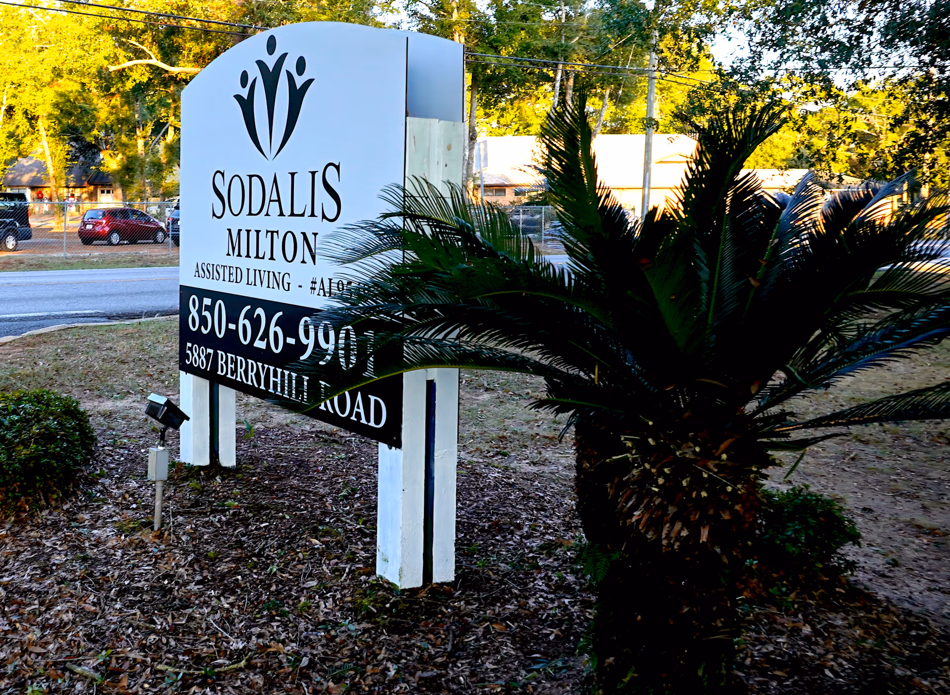 Outdoor sign for Sodalis Milton Assisted Living located at 5887 Berryhill Road, with a phone number 850-626-9901, surrounded by trees and plants near a road.