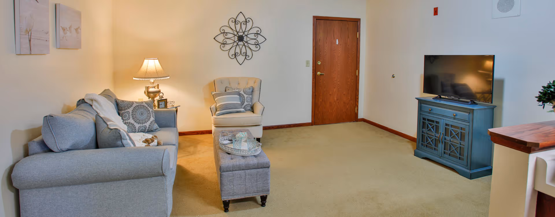 A cozy living room with a gray sofa adorned with patterned pillows and a throw blanket, a beige armchair with cushions, a small ottoman with a decorative tray, a table lamp on a side table, a flat-screen TV on a blue cabinet, and wall decorations including two framed pictures and a metal wall art piece. The room has beige carpet and light-colored walls.