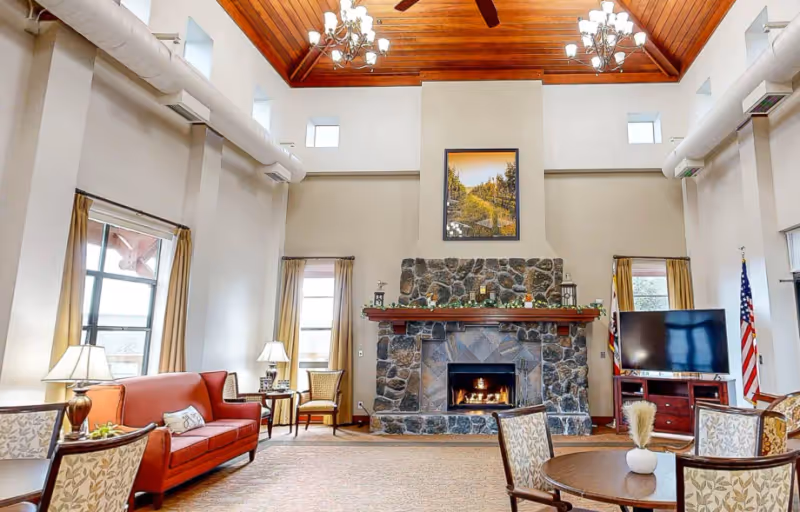 A spacious living room with a high wooden ceiling featuring chandeliers and ceiling fans. The room has a large stone fireplace with a fire burning and a framed picture above it. There are multiple windows with curtains, a red sofa, several armchairs, a round wooden table with a small vase, lamps, and a flat-screen TV on a wooden stand. An American flag is visible in the corner.