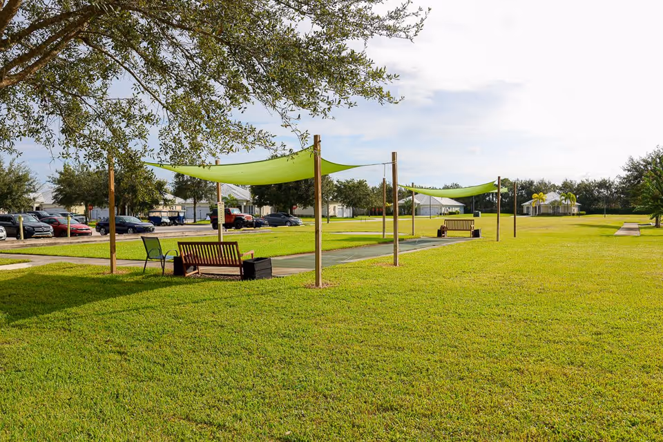 A green outdoor lawn area with two shaded benches and a bocce ball court. There are trees, parked cars, and residential buildings in the background under a partly cloudy sky.
